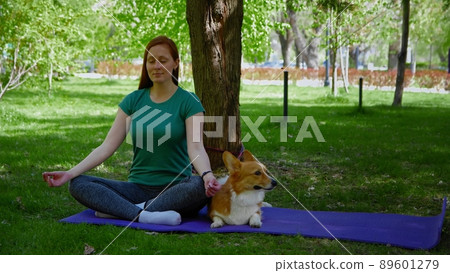 A young woman meditates in the park with her corgi dog. Outdoor sports A young woman meditates in the park with her corgi dog. Outdoor sports 89601279