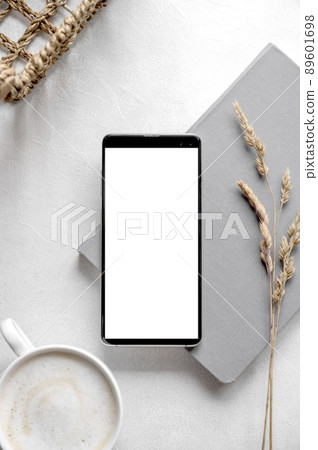 Modern tabletop with with smartphone mockup, cup of coffee, dried plants and notebook on textured white background. Flat lay, minimal style workspace 89601698
