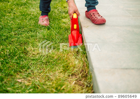 Close up of man hand with red hedge trimmer cutting green grass at spring time Close up of man hand with red hedge trimmer cutting green grass at spring time 89605649