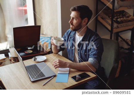 Thoughtful Caucasian handsome man drinking coffee in cafe and working at laptop. Coffee break. 89609581