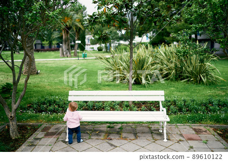 Little girl stands near a white bench in the park Little girl stands near a white bench in the park 89610122