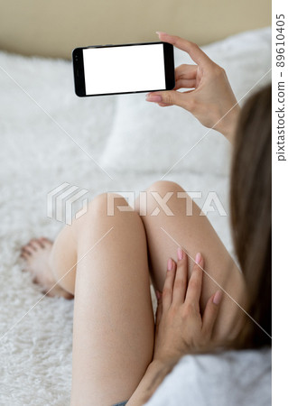 Online meeting. Mobile mockup. Virtual connection. Unrecognizable relaxed woman looking smartphone with blank screen sitting bed in light room interior. Online meeting. Mobile mockup. Virtual connection. Unrecognizable relaxed woman looking smartphone with blank screen sitting bed in light room interior. 89610405