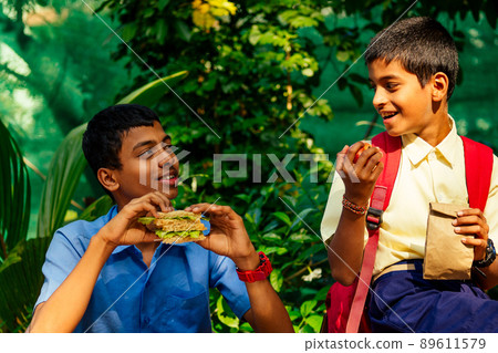 indian school boy eating sandwich and his friend with backpack sits on table and eating an apple in park 89611579