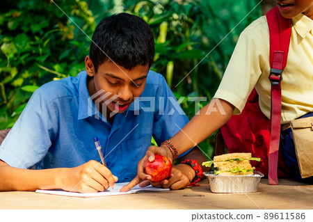 indian school boy writing on notepad doing homework , looking concentration . His friend with backpack sits on table and eating an apple in park indian school boy writing on notepad doing homework , looking concentration . His friend with backpack sits on table and eating an apple in park 89611586