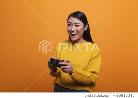 Beautiful cheerful excited young adult woman playing video games wtih modern controller while enjoying leisure time. Playful positive person with gaming gamepad wearing yellow sweater. 89612313
