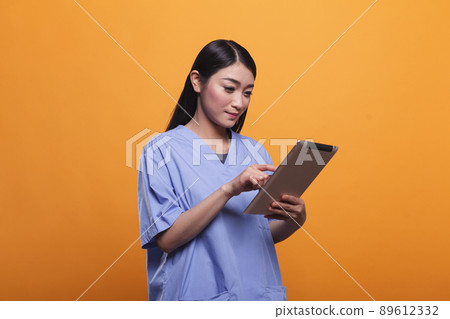 Pretty asian hospital nurse wearing blue medical uniform while using modern tablet on orange background. Clinic healthcare staff member using digital tablet. Studio shoot. Pretty asian hospital nurse wearing blue medical uniform while using modern tablet on orange background. Clinic healthcare staff member using digital tablet. Studio shoot. 89612332