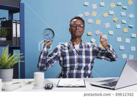 Development company office smiling worker congratulating coworker by showing thumbs up symbol while having clock. African american agency employee showing excellent job gesture while in workspace. 89612382
