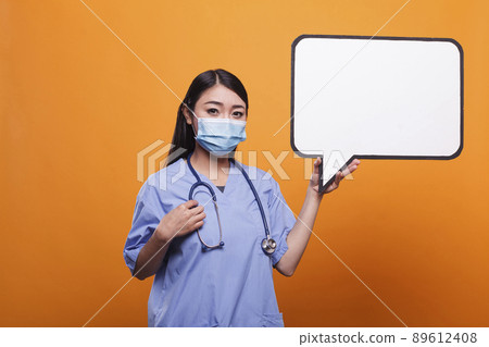 Confident young adult healthcare clinic asian nurse wearing facemask and blue uniform while holding cardboard speech bubble. Hospital caregiver holding cardboard speech bubble sign. 89612408