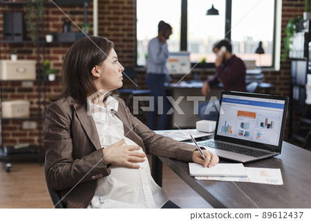 Pregnant businesswoman at office desk using laptop to analyze data charts and project management. Expectant mother reviewing company annual results while sitting in agency workplace. 89612437