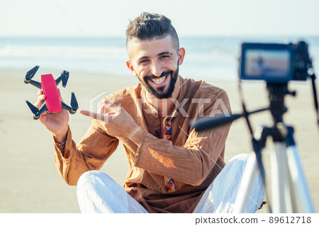 arabian man sitting on sand and talking tripod video making review of quadrocopter in beach. Travel videographer lifestyle content maker concept 89612718