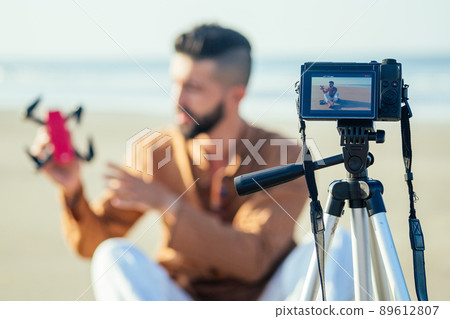arabian man sitting on sand and talking tripod video making review of quadrocopter in beach. Travel videographer lifestyle content maker concept arabian man sitting on sand and talking tripod video making review of quadrocopter in beach. Travel videographer lifestyle content maker concept 89612807