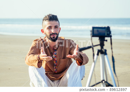 arabian man sitting on sand and talking tripod video making in beach. Travel videographer lifestyle content maker concept 89612897