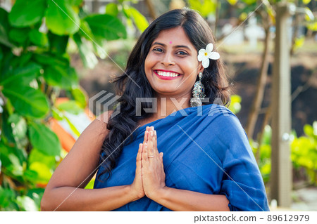 cheerful young indian woman with white flover in hair relaxing hand gesture namaste on beach in Goa 89612979