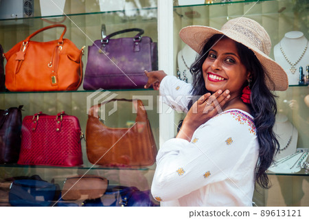 happy indian woman wearing straw hat and standind near showcase with fashion bags in India Goa market happy indian woman wearing straw hat and standind near showcase with fashion bags in India Goa market 89613121