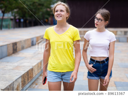 Portrait of two positive girls walking along street of summer city Portrait of two positive girls walking along street of summer city 89613367