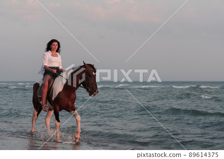 Woman in summer clothes enjoys riding a horse on a beautiful sandy beach at sunset. Selective focus Woman in summer clothes enjoys riding a horse on a beautiful sandy beach at sunset. Selective focus 89614375