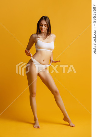 Portrait of young beautiful woman with slim figure posing in white underwear isolated over yellow studio background. Depilation, epilation Portrait of young beautiful woman with slim figure posing in white underwear isolated over yellow studio background. Depilation, epilation 89618697