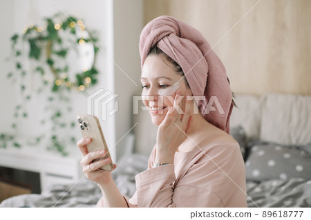Woman in pajama with eyes patches and towel on head using phone in bed. Morning habits and home beauty routine Woman in pajama with eyes patches and towel on head using phone in bed. Morning habits and home beauty routine 89618777