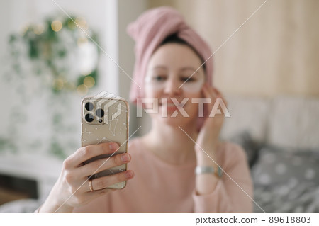 Woman in pajama with eyes patches and towel on head using phone in bed. Morning habits and home beauty routine 89618803