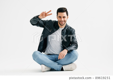 Positive cheerful young man doing victory sign sitting with legs crossed on floor isolated over white background Positive cheerful young man doing victory sign sitting with legs crossed on floor isolated over white background 89619231