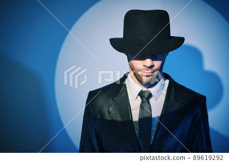 Portrait of handsome man in black coat hiding face behind his hat in the spotlight on studio background Portrait of handsome man in black coat hiding face behind his hat in the spotlight on studio background 89619292
