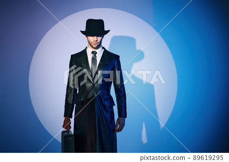 Portrait of handsome man in black coat hiding face behind his hat in the spotlight on studio background 89619295