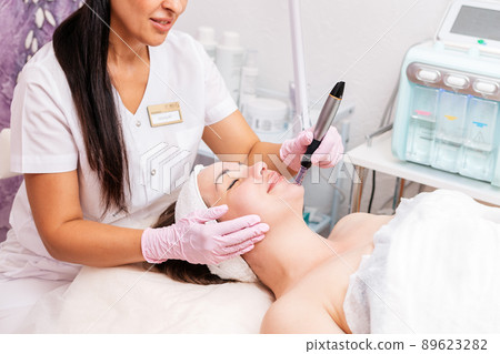 Beauty salon. Smiling cosmetologist in rubber gloves makes fractional mesotherapy of the face to the client. Close up. Concept of professional modern cosmetology Beauty salon. Smiling cosmetologist in rubber gloves makes fractional mesotherapy of the face to the client. Close up. Concept of professional modern cosmetology 89623282
