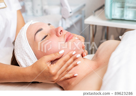Cosmetologist makes a woman a facial massage procedure. Portrait of the client and the doctor's hands in close-up. Concept of cosmetology and beauty Cosmetologist makes a woman a facial massage procedure. Portrait of the client and the doctor's hands in close-up. Concept of cosmetology and beauty 89623286
