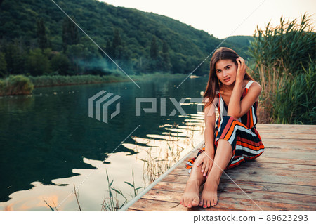A young pretty woman poses sitting on a wooden pier by the river. Summer holiday A young pretty woman poses sitting on a wooden pier by the river. Summer holiday 89623293