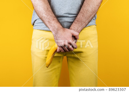 Impotence and men's health. A man in yellow jeans holds a banana in his hands, covering his genitals. Hands and banana close up. Yellow background 89623307