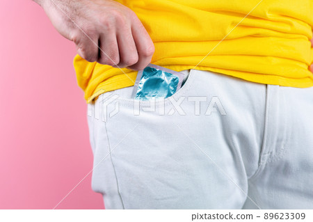 A man in white jeans pulls a condom out of his pocket. Close up. Pink background. Concept of protection against sexually transmitted infections A man in white jeans pulls a condom out of his pocket. Close up. Pink background. Concept of protection against sexually transmitted infections 89623309