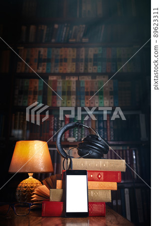 Grouped Mobile phone, headphones and a stack of books near the lamp on a wooden table. Concept of training and audiobooks. Library bookcase in the background with light. Copy space. Mock up Grouped Mobile phone, headphones and a stack of books near the lamp on a wooden table. Concept of training and audiobooks. Library bookcase in the background with light. Copy space. Mock up 89623311