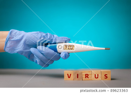 A hand in a blue medical glove holds an electronic thermometer with the inscription FLU. At hand are wooden cubes with the inscription virus.The concept of coronavirus and symptoms of the disease 89623363