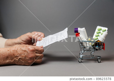 Close up of hands of senior woman holding an instructions of medicines near shopping cart. Gray background. Copy space. Concept of shopping pills and treatment Close up of hands of senior woman holding an instructions of medicines near shopping cart. Gray background. Copy space. Concept of shopping pills and treatment 89623378
