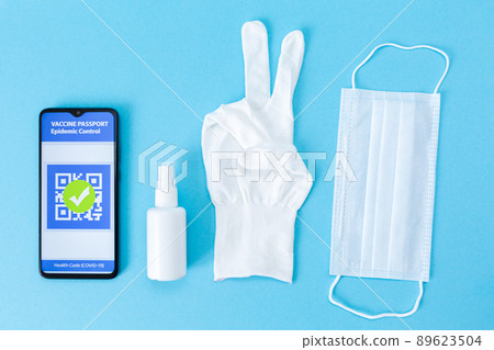 An electronic passport of immunity with a mark on the COVID-19 vaccination on the cellphone screen, antiseptic, protective mask and white glove showing a gesture of victory. Flat lay An electronic passport of immunity with a mark on the COVID-19 vaccination on the cellphone screen, antiseptic, protective mask and white glove showing a gesture of victory. Flat lay 89623504