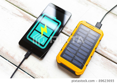 Concept of modern technologies and power bank. Smartphone with a turquoise battery icon, charged from a portable solar charger. White wooden background with stripes. Close up Concept of modern technologies and power bank. Smartphone with a turquoise battery icon, charged from a portable solar charger. White wooden background with stripes. Close up 89623693