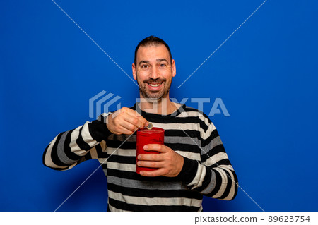 Smiling Latin man with a beard dressed in a striped sweater putting a euro coin in a metal piggy bank, trying to save so he can travel on his vacation. Isolated on blue studio background Smiling Latin man with a beard dressed in a striped sweater putting a euro coin in a metal piggy bank, trying to save so he can travel on his vacation. Isolated on blue studio background 89623754