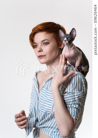 Sphynx Hairless Cat sitting on shoulder of redhead young woman. Hipster female with short hair dressed in striped white-blue shirt. Studio shot on white background. Part of series. Selective focus 89626464