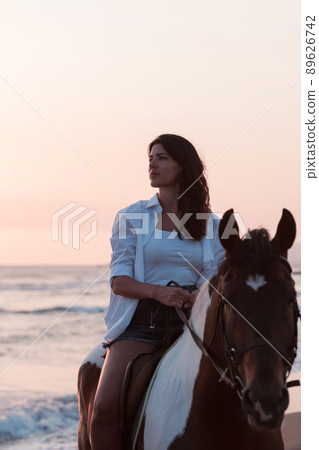 Woman in summer clothes enjoys riding a horse on a beautiful sandy beach at sunset. Selective focus Woman in summer clothes enjoys riding a horse on a beautiful sandy beach at sunset. Selective focus 89626742
