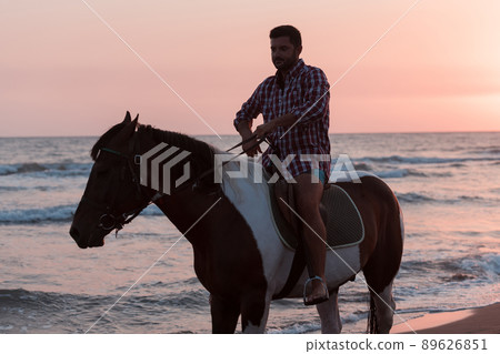 A modern man in summer clothes enjoys riding a horse on a beautiful sandy beach at sunset. Selective focus A modern man in summer clothes enjoys riding a horse on a beautiful sandy beach at sunset. Selective focus 89626851