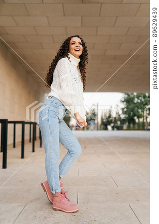 Girl in urban dress with dark hair curls cheerfully smiles at someone in the distance Girl in urban dress with dark hair curls cheerfully smiles at someone in the distance 89629439
