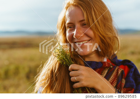 A girl without makeup with red hair smiles broadly and holds a bouquet of wild flowers in her hand against the background of the steppe and hills. close-up 89629585