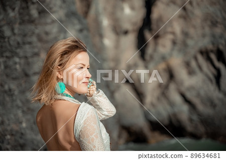 Middle aged woman looks good with blond hair, boho style in white long dress on the beach decorations on her neck and arms. 89634681
