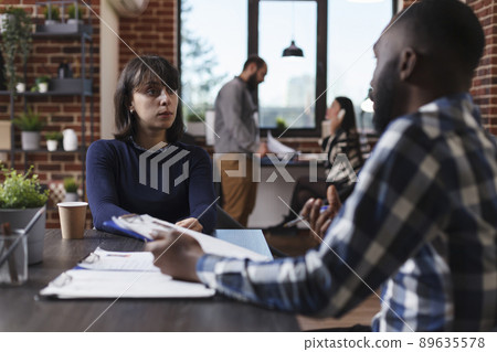 Business company recruiter and candidate sitting at desk in office while reviewing job applicant CV. Multiethnic people in startup agency talking about salary offer and internal regulations. 89635578