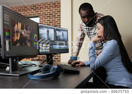 Post production house employee talking with coworker about footage quality. Professional movie editor sitting at desk while improving film frames using advanced specialized software. Post production house employee talking with coworker about footage quality. Professional movie editor sitting at desk while improving film frames using advanced specialized software. 89635750