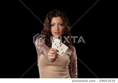 Brunette girl wearing shiny dress posing holding two playing cards in her hand standing against black studio background. Casino, poker. Close-up. 89642639