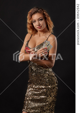 Ginger girl in shiny dress posing holding two playing cards and chips in her hands standing against black studio background. Casino, poker. Close-up. 89642642