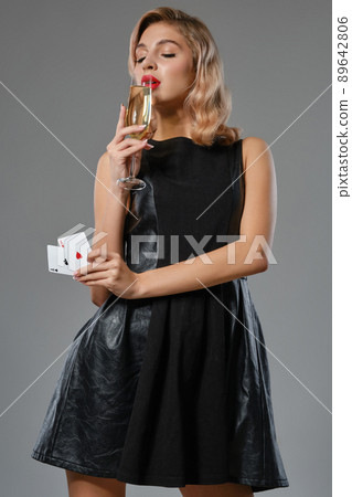 Blonde girl in black dress showing two playing cards, drinking champagne, posing against gray studio background. Gambling, poker, casino. Close-up. Blonde girl in black dress showing two playing cards, drinking champagne, posing against gray studio background. Gambling, poker, casino. Close-up. 89642806