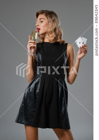 Blonde girl in black dress showing two playing cards, drinking champagne, posing against gray studio background. Gambling, poker, casino. Close-up. 89642845