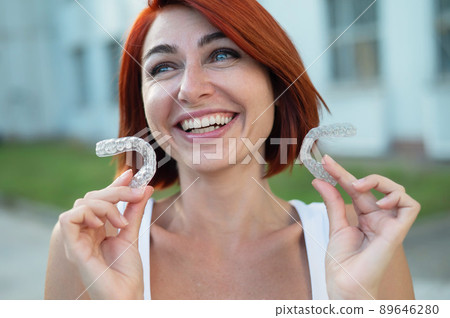 Red-haired Caucasian woman holding transparent mouthguards for bite correction outdoors. A girl with a beautiful snow-white smile uses silicone braces Red-haired Caucasian woman holding transparent mouthguards for bite correction outdoors. A girl with a beautiful snow-white smile uses silicone braces 89646280